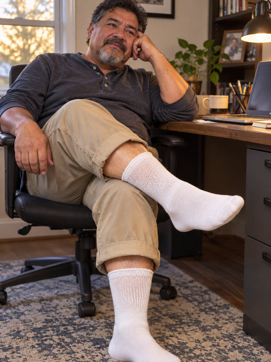 Man seated at a home office desk with one ankle over his knee, wearing white non-binding diabetic crew socks